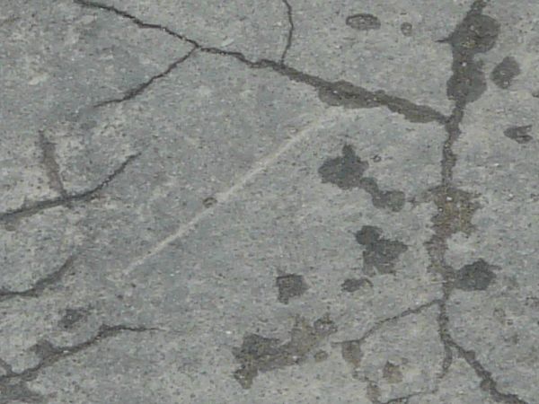 Grey asphalt texture, covered with cracks of various sizes and depths, and many shallow chipped holes.
